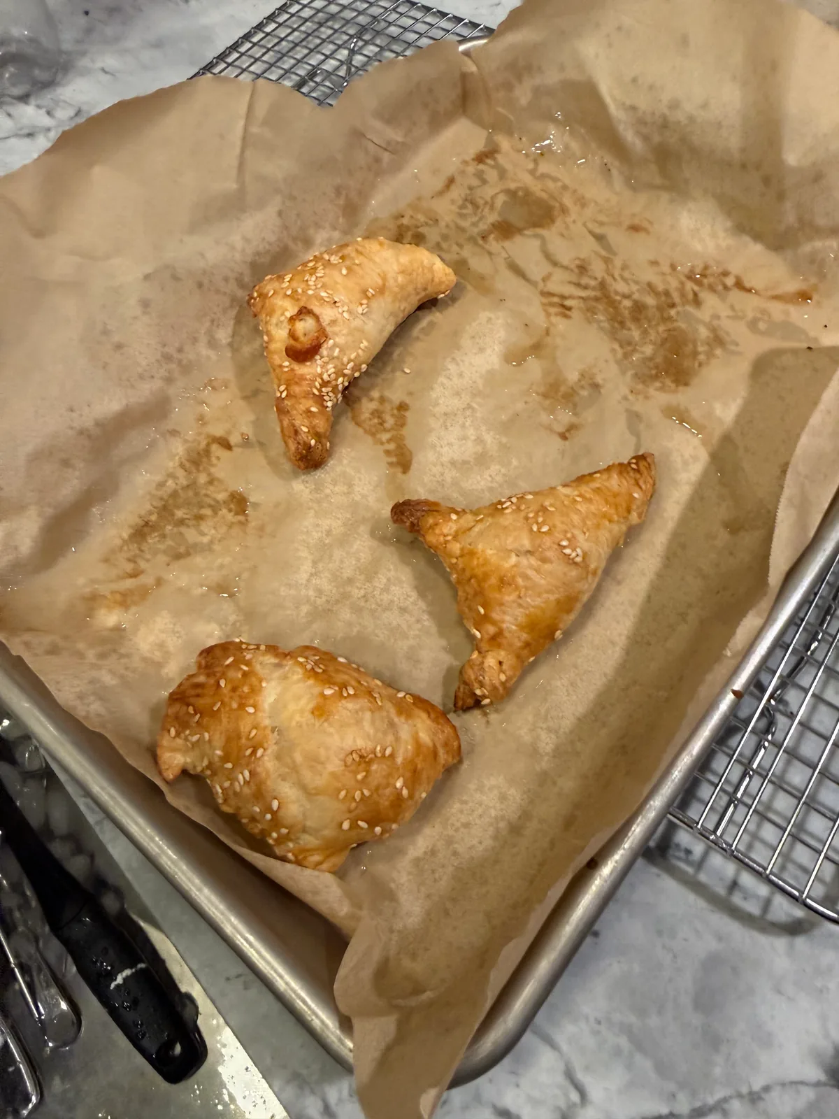 Three golden-brown, sesame-topped pastries—two triangular and one rounded—rest on grease-stained parchment paper in a metal baking sheet.