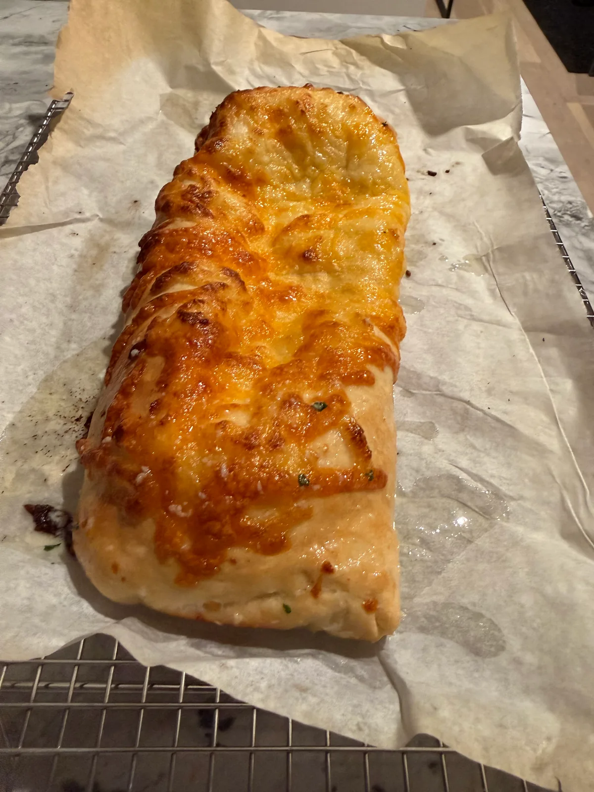 Long rectangular loaf of baked bread topped with melted, golden-brown cheese, resting on parchment over a wire rack.