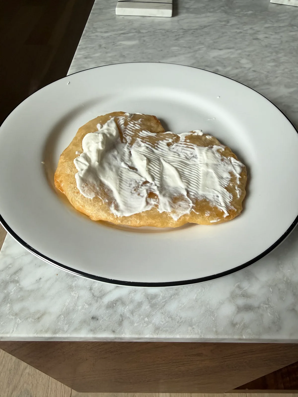 A round, golden-brown flatbread covered with a thick white spread sits on a white plate with a thin black rim on a marble-patterned table.