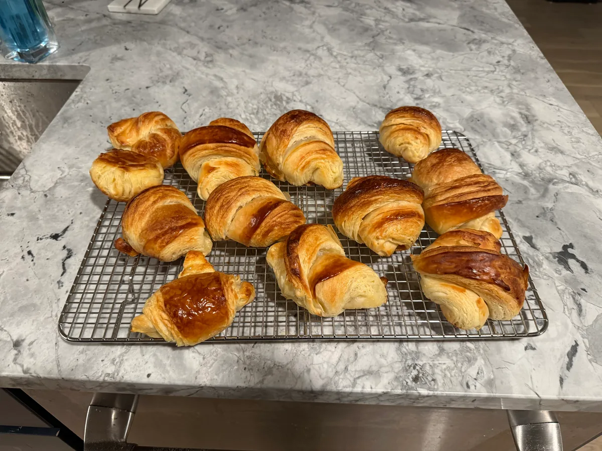 About a dozen golden-brown, flaky croissants of varying shapes and sizes cooling on a rectangular wire rack on a gray marble countertop.
