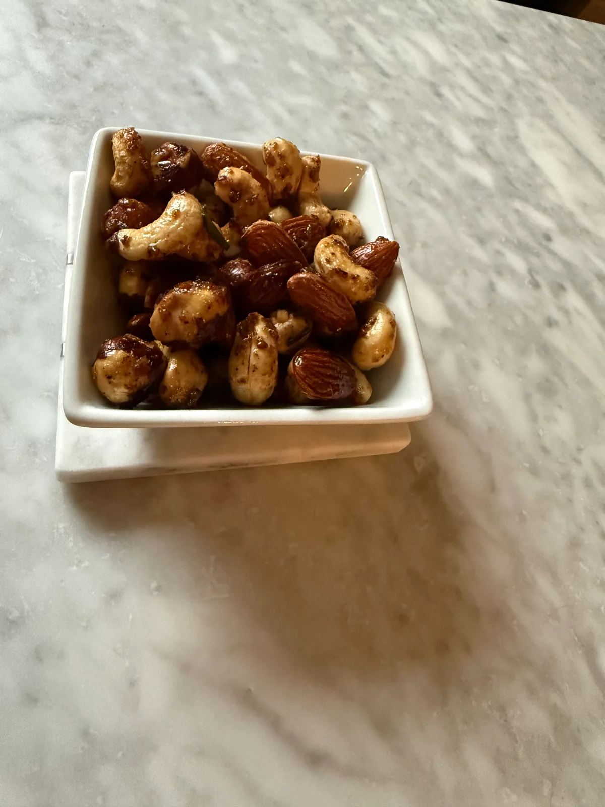A small square white bowl filled with glazed mixed nuts, including almonds and cashews, on a white saucer atop a marble table.