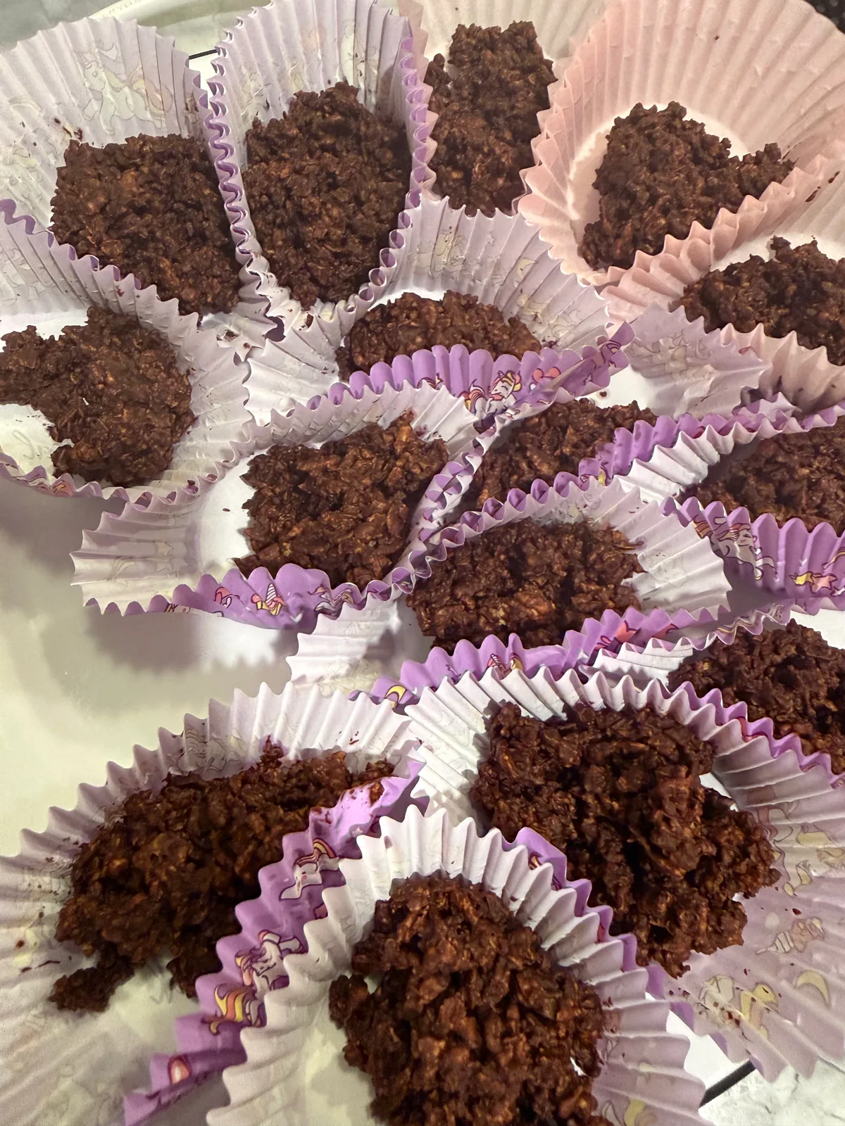Rough dark chocolate-coated flake clusters sit in opened white, pink, and purple pleated paper cups, crowded together on a light surface.