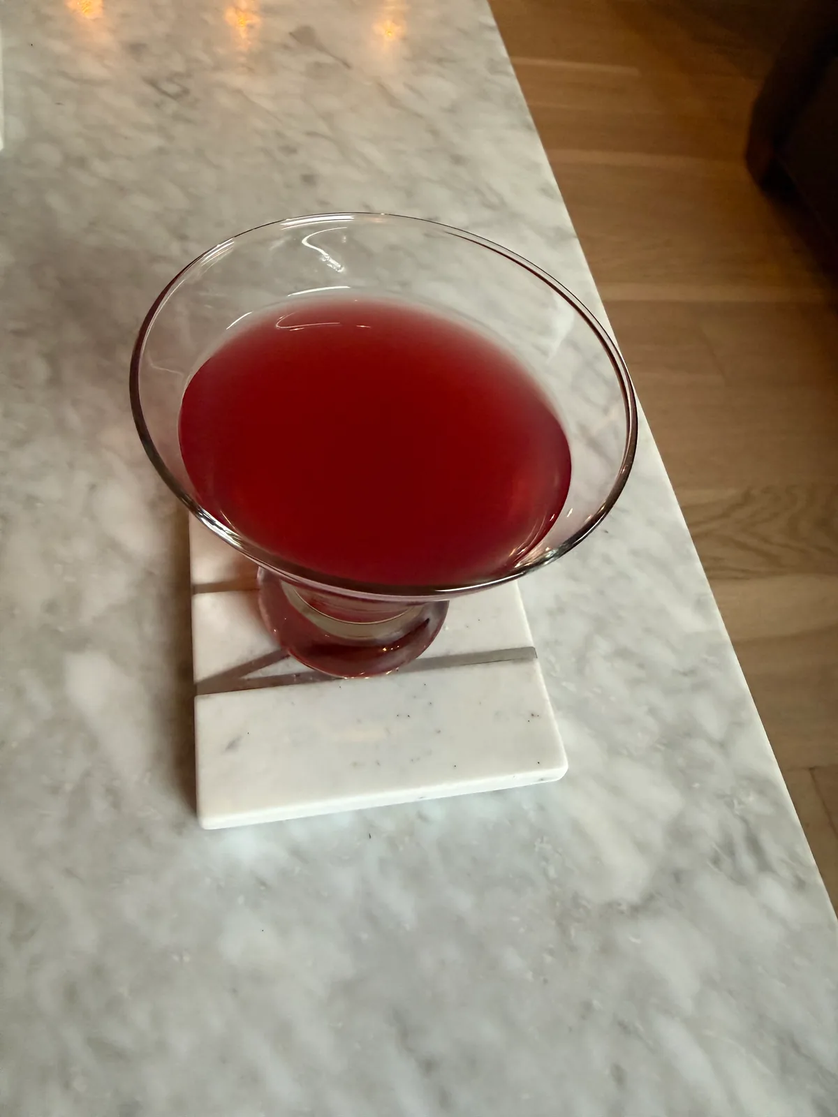 A clear, stemmed cocktail glass filled with a deep red drink sits on a square white coaster atop a gray marble counter.