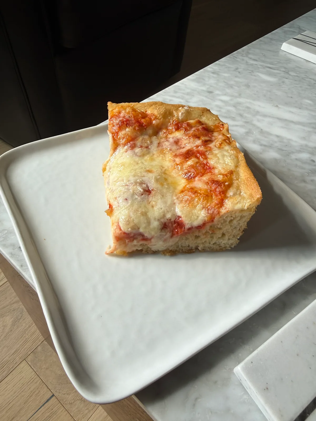 A thick square slice of pan pizza with browned melted cheese and patches of red tomato sauce sits on a white rectangular plate on a marble countertop.