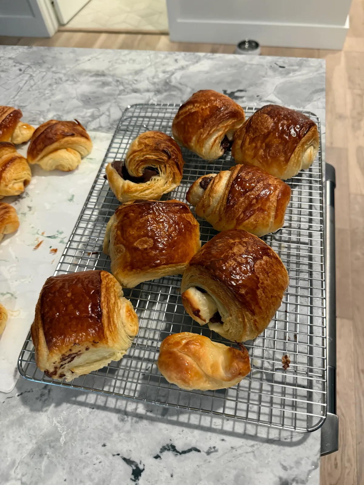 Several deeply browned, flaky laminated pastries cooling on a rectangular wire rack on a marble-patterned countertop; some are rectangular with dark chocolate visible at the ends, with additional pastries partly visible at left.
