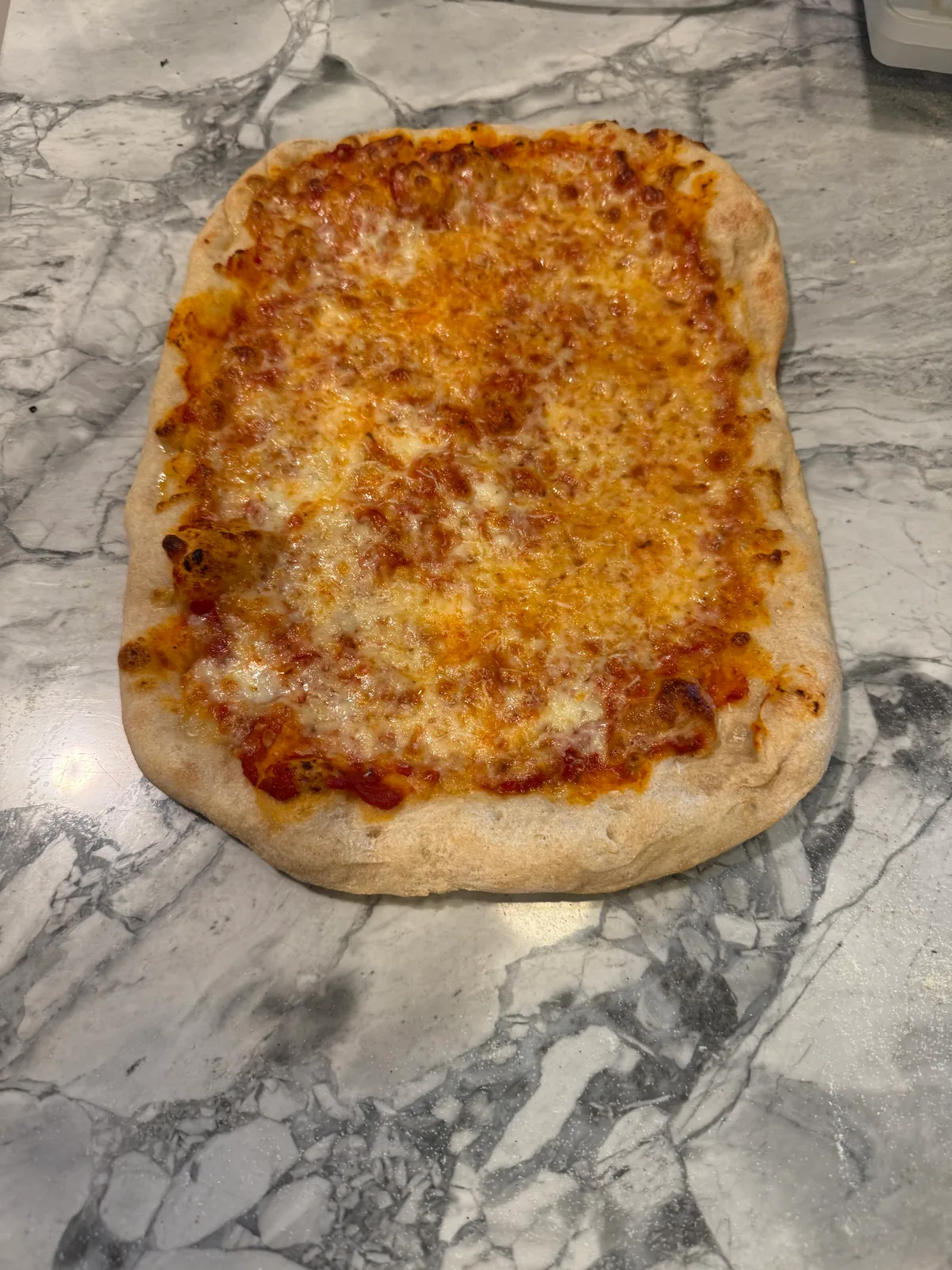 Rectangular homemade cheese pizza with browned melted cheese, visible tomato sauce, and a thick pale-golden crust on a gray marble counter.
