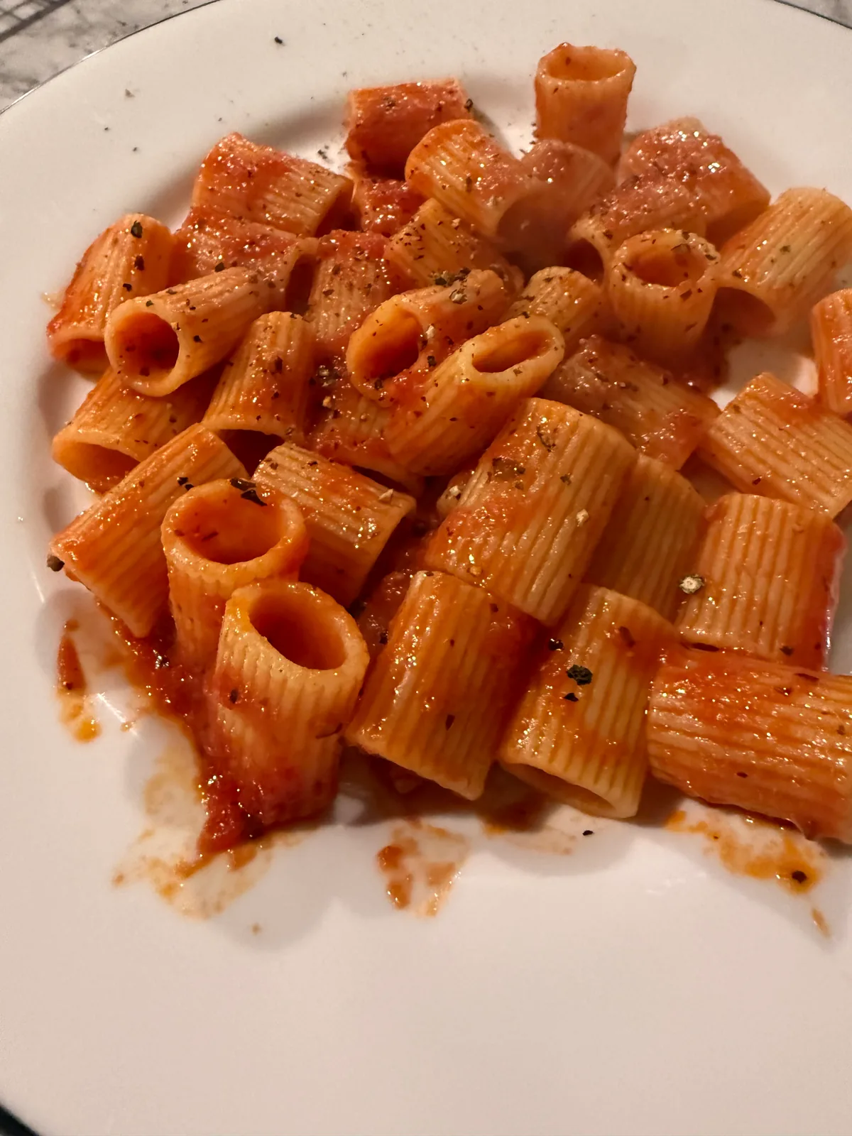 Tube-shaped ridged pasta coated in red tomato sauce, sprinkled with black pepper, on a white plate.
