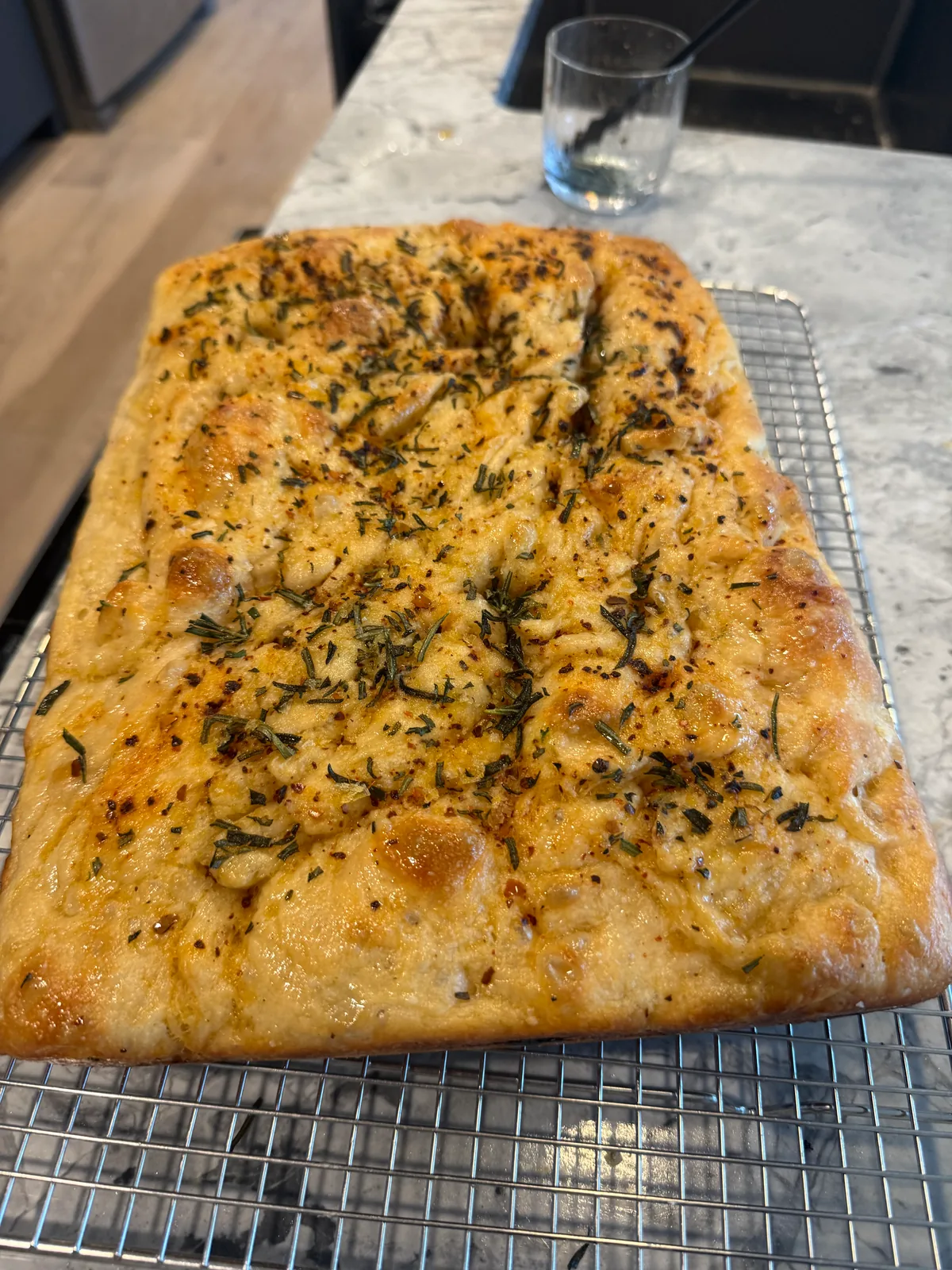Rectangular golden bread with a glossy, dimpled surface scattered with herbs, resting on a wire cooling rack on a kitchen counter.