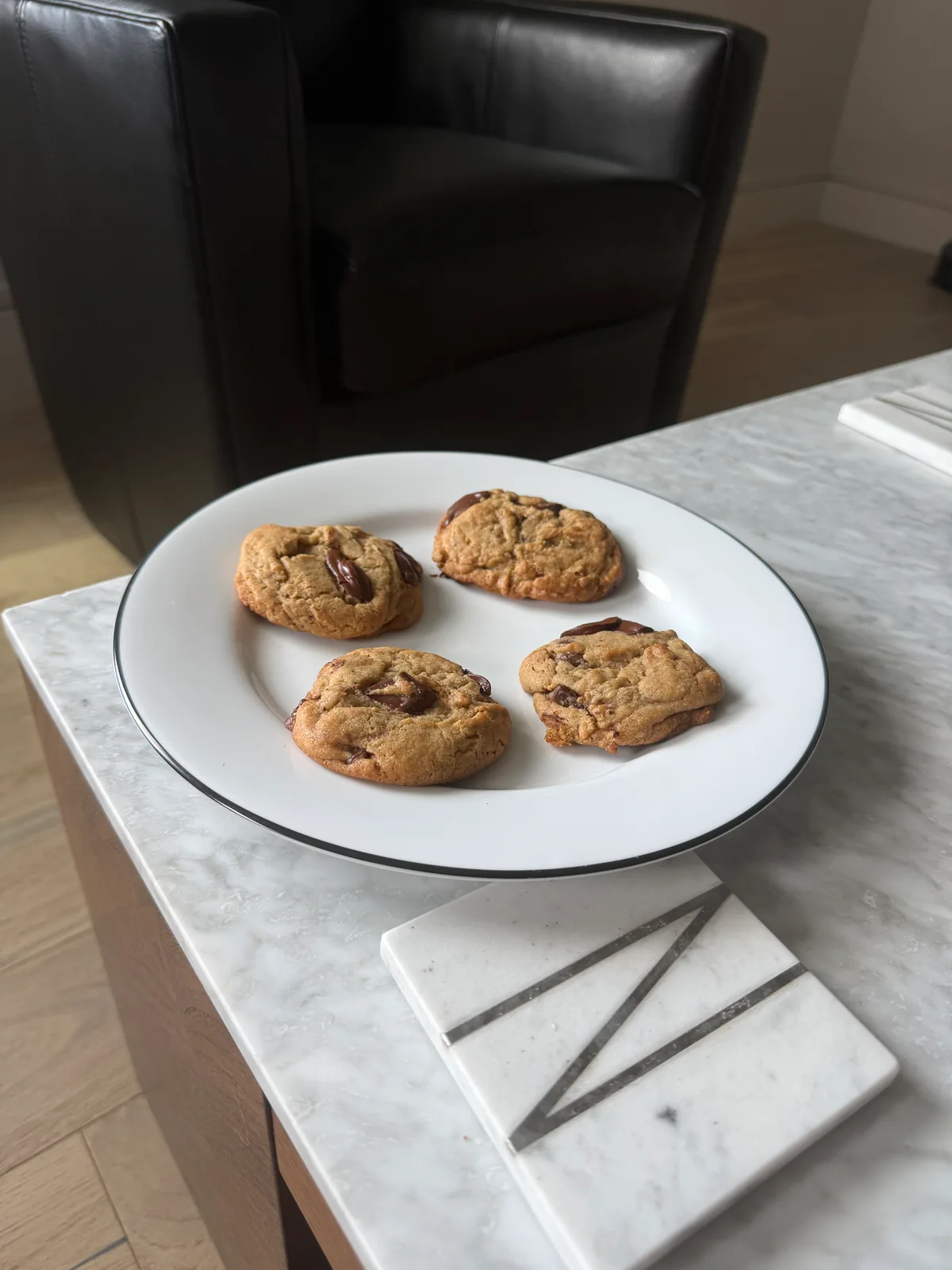 Four golden-brown chocolate chunk cookies arranged on a white plate with a thin dark rim, set on a light marble table beside a square marble coaster.
