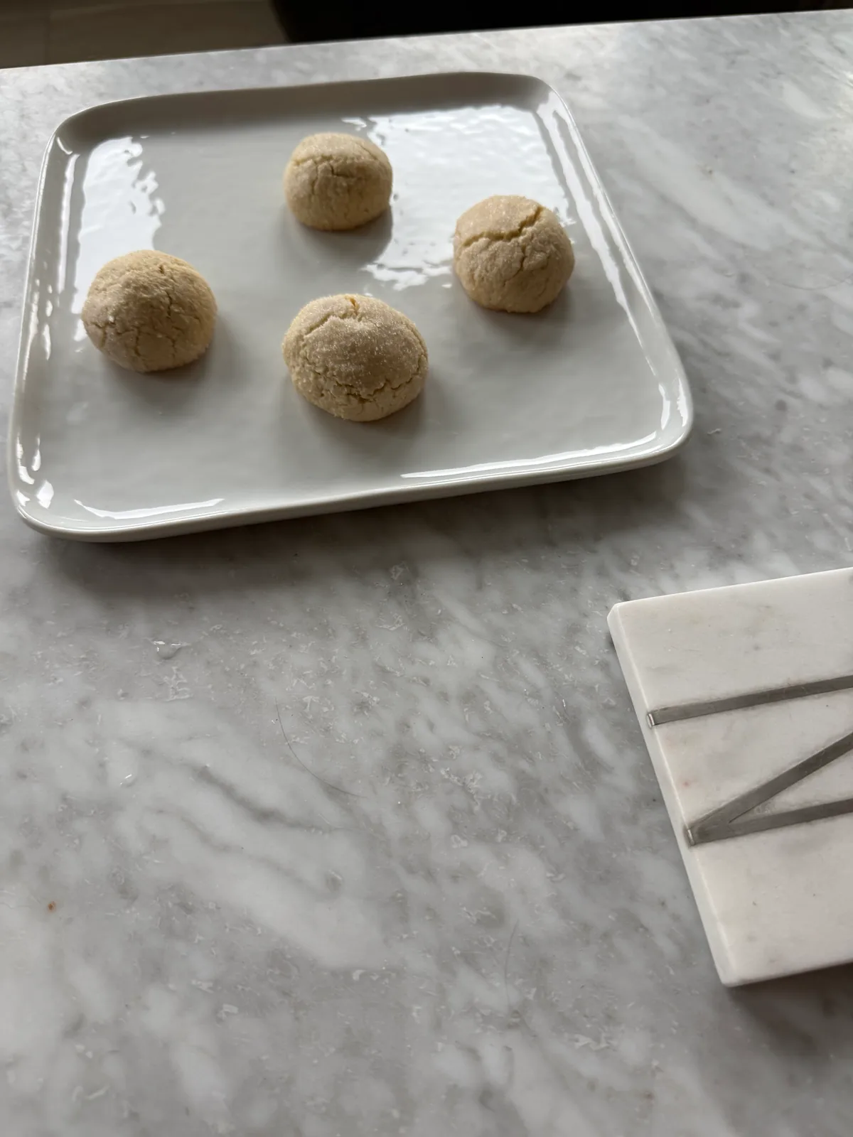 Four pale, round, sugar-coated cookies with cracked surfaces sit on a white rectangular plate on a marble countertop.