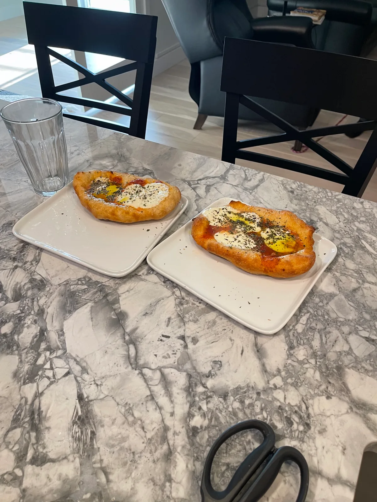 Two small oval fried dough pizzas topped with tomato sauce, white cheese, and dried herbs sit on separate square white plates on a gray marble-patterned table; an empty drinking glass and black dining chairs are nearby.